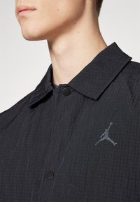 Homme portant une veste noire texturée avec des boutons-pression et un logo Jumpman réfléchissant sur la poitrine, visible du cou jusqu'au milieu du torse.