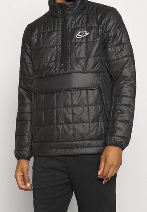 Homme portant une veste Nike noire matelassée à enfiler avec une demi-fermeture éclair avant et une grande poche avant.