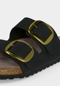 Μαύρο υφασμάτινο σανδάλι Birkenstock με δύο ρυθμιζόμενες χρυσές αγκράφες και φελό πάτο σε ανοιχτό φόντο.