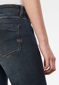 Jeans de mezclilla azul oscuro con un diseño de cintura alta, que cuentan con un gran bolsillo trasero y detalles de costura sutiles sobre una textura suave.