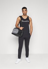 Camiseta sin mangas negra de Champion y pantalones de chándal. Lleva una bolsa de deporte negra y gris con textura de malla. Zapatillas blancas. Fondo simple.