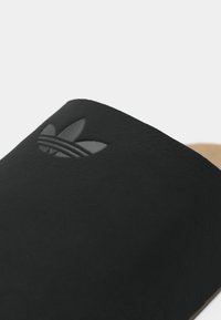 Melnas slīdvirsmas sandales ar gludu ādas virsu, kas piedāvā debosētu logotipu. Iekšpusē ir brūnas, strukturētas zoles, kas nodrošina komfortu.