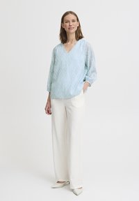 Blusa a fantasia blu chiaro con scollatura a V e maniche a tre quarti, abbinata a pantaloni bianchi a gamba larga e ballerine crema.