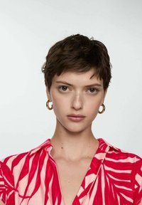 Tričko s krátkym rukávom s ružovým pozadím a červenými listovými vzormi. Modelka má na sebe zlaté kruhové náušnice a prirodzený make-up.