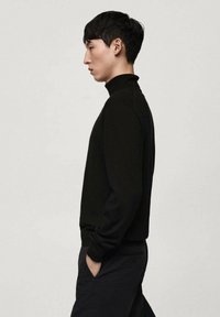 Svart polotröja i mjukt, texturerat stickat material, med långa ärmar och en figurnära siluett, som bärs med mörka byxor.