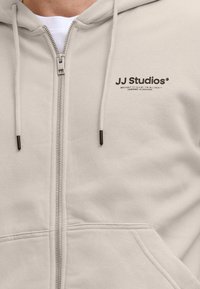 Helle beige Kapuzenjacke mit Reißverschluss aus weichem Stoff, ausgestattet mit einem Logoprint und Kordeln mit Metallenden, sichtbaren Nähten und einer Vordertasche.