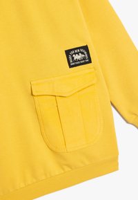 Sudadera amarilla con un gran bolsillo frontal con tapa, puños acanalados y una pequeña etiqueta de marca con texto negro y detalle gráfico.