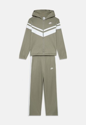 Olivovo zelený Nike tréningový komplet, zapínacia bunda s bielymi chevronovými pruhmi a zodpovedajúcimi nohavicami. Má kapucňu a vyšívané logo na hrudi a stehne.