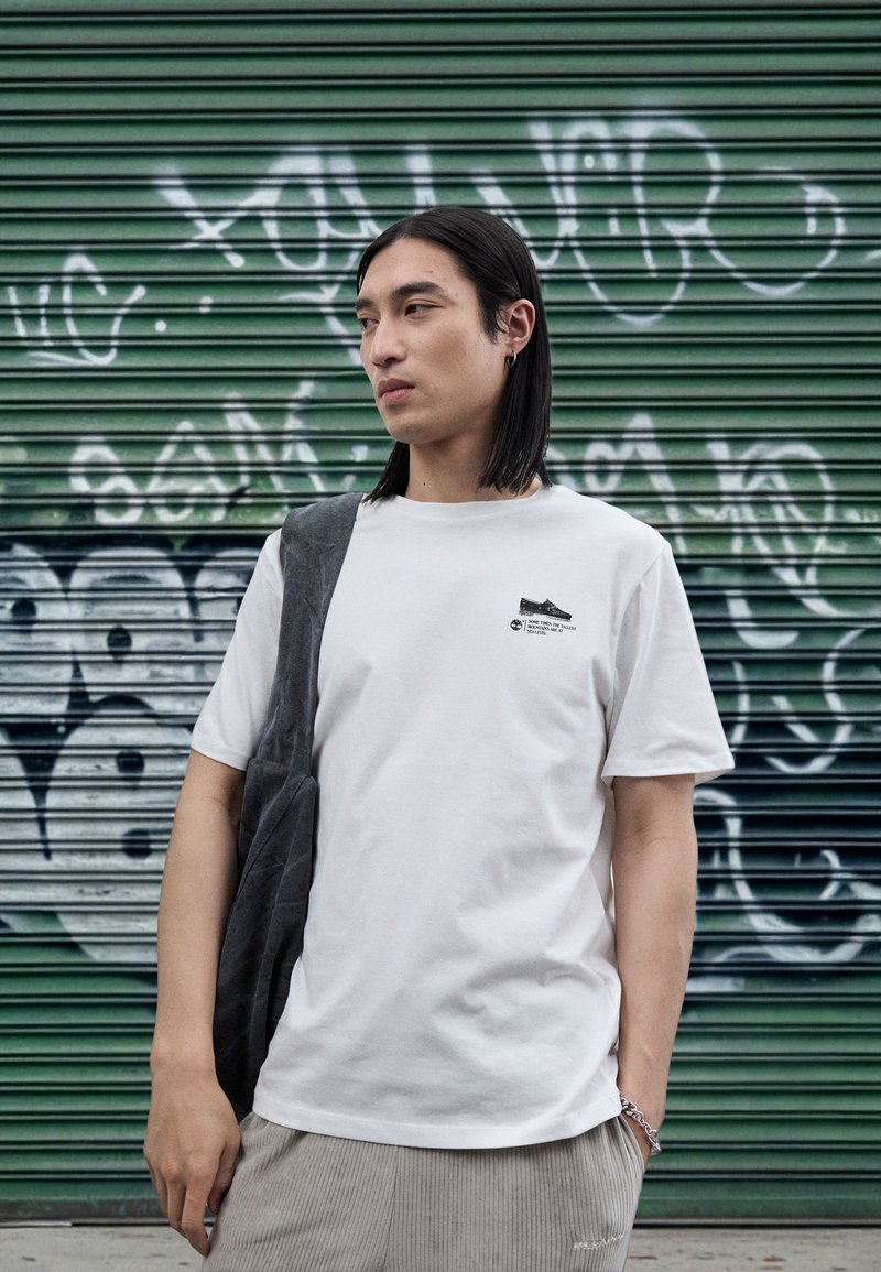 White cotton t-shirt with a graphic print on the chest, paired with gray wide-leg corduroy pants and a gray fabric sling. Green graffiti backdrop.