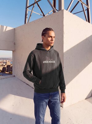 Un homme portant un sweat à capuche foncé et un jean bleu se tient au soleil sur une terrasse sur le toit, avec une vue sur la ville et des murs en béton derrière lui.