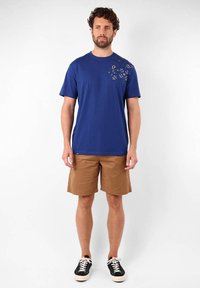 T-shirt en coton bleu avec un col rond et des manches courtes, présentant un imprimé décoratif sur la poitrine gauche. Associée à un short marron et des baskets noires.