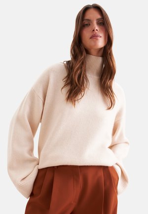 Femme aux longs cheveux bruns portant un pull à col roulé beige et un pantalon taille haute de couleur rouille, les mains dans les poches, sur fond uni.