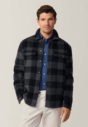 Chaqueta de franela a cuadros en negro y gris, con dos bolsillos en el pecho, cierre de botones y cuello. Llevada sobre una camisa de mezclilla azul.
