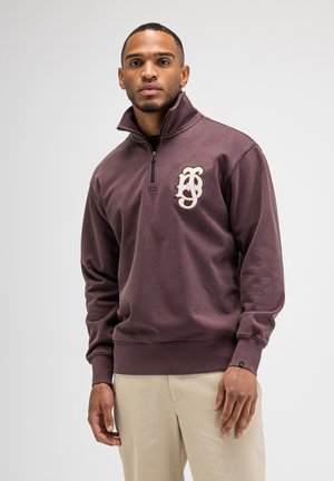 Mann trägt einen dunkelvioletten Pullover mit Viertel-Reißverschluss und weißem Logo auf der Brust sowie beigefarbene Hosen vor einem einfarbigen hellen Hintergrund.