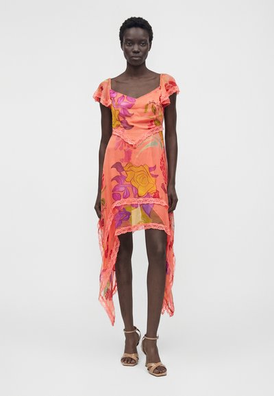 Γυναίκα φοράει κοραλλί ροζ φόρεμα με υψηλή-χαμηλή γραμμή, διακοσμημένο με δαντέλα και μεγάλα λουλουδάτα σχέδια σε κίτρινο, μωβ και κόκκινο, συνδυασμένο με μπεζ τακούνια.