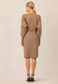 Geripptes, beiges Strickkleid mit langen Ärmeln, tailliertem Schnitt und knielangem Rock. Kombiniert mit schwarzen, verzierten Absatzschuhen. Rückansicht, glatte Textur.