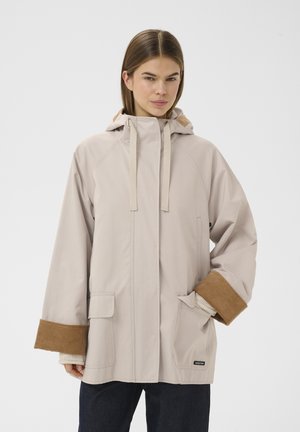 Giovane donna con giacca beige con cappuccio e polsini in velluto a coste marrone, in piedi con una mano in tasca contro uno sfondo bianco uniforme.