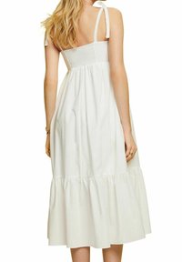 Femme aux longs cheveux blonds portant une robe midi blanche sans manches avec un corsage smocké et des bretelles nouées sur les épaules, vue de dos.