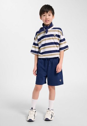 Dreng står, iført marineblå shorts, stribet beige og marineblå t-shirt, hvide sneakers, hvide sokker og et marineblåt tørklæde.