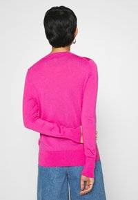 Pull en maille rose à manches longues et poignets côtelés. Le dos présente un design sans coutures et une silhouette légèrement ajustée, complétés par un jean bleu.