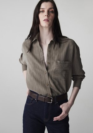 Młoda osoba z długimi ciemnymi włosami nosi luźną oliwkową koszulę i ciemne dżinsy, stojąc z jedną ręką w kieszeni na jednolitym tle.