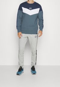 Sudadera en verde oscuro y blanco con un diseño de chevrones; combinada con pantalones de chándal grises que presentan tiras laterales negras y detalles de logotipo.