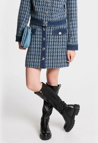 Vestido de tweed con estampado azul y acentos de denim, con botones en el frente y dos bolsillos laterales; combinado con botas de piel negra hasta la rodilla con suelas texturizadas.