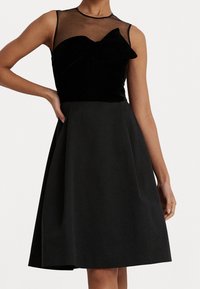 Svart klänning med velouröverdelen och ett genomskinligt ok, en rosett-detajl och en utsvängd kjol. Slät textur, ärmlös design, knälång.