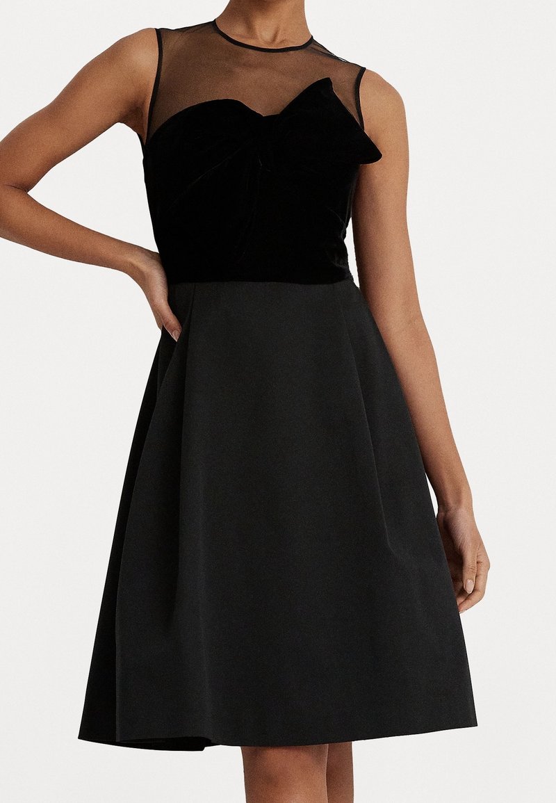 Svart klänning med velouröverdelen och ett genomskinligt ok, en rosett-detajl och en utsvängd kjol. Slät textur, ärmlös design, knälång.