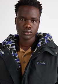 Schwarze Jacke mit gemustertem Fleece-Futter in Blau- und Cremetönen, mit einem Logo-Patch auf der Brust und einem Hemd mit Kragen darunter.