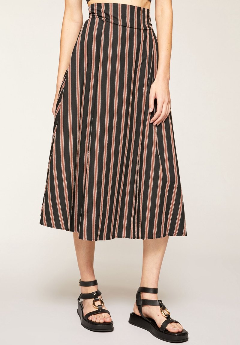 Jupe noire taille haute avec des rayures verticales rouges et blanches portée avec des sandales noires à bout ouvert avec des détails en anneaux dorés.