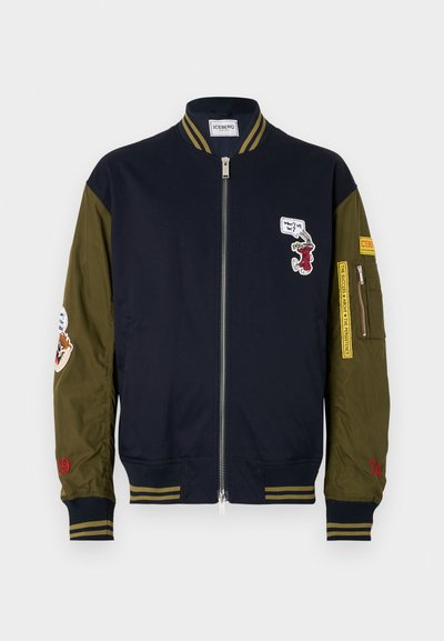 Marineblaues und olivgrünes Bomberjackett mit gerippten Bündchen, Reißverschluss, gestickten Patches und Akzentnähten in Rot und Gelb.
