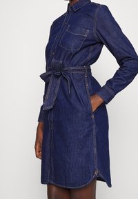 Denimskjortklänning i mörkblått med långa ärmar, knäppning, sidofickor och midjeband. Har en enkel br östficka och kontrasterande sömmar.