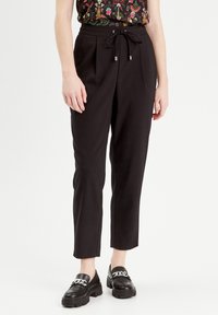 Pantalon noir avec taille à cordon, plis et jambes fuselées. Porté avec des mocassins noirs présentant un détail en chaîne argentée.