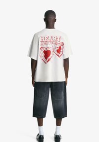 Tricou alb cu mânecă scurtă și inscripția roșie "HEART Struck" împreună cu două graficuri în formă de inimă pe spate, asortat cu pantaloni șort din denim închis și pantofi negri.