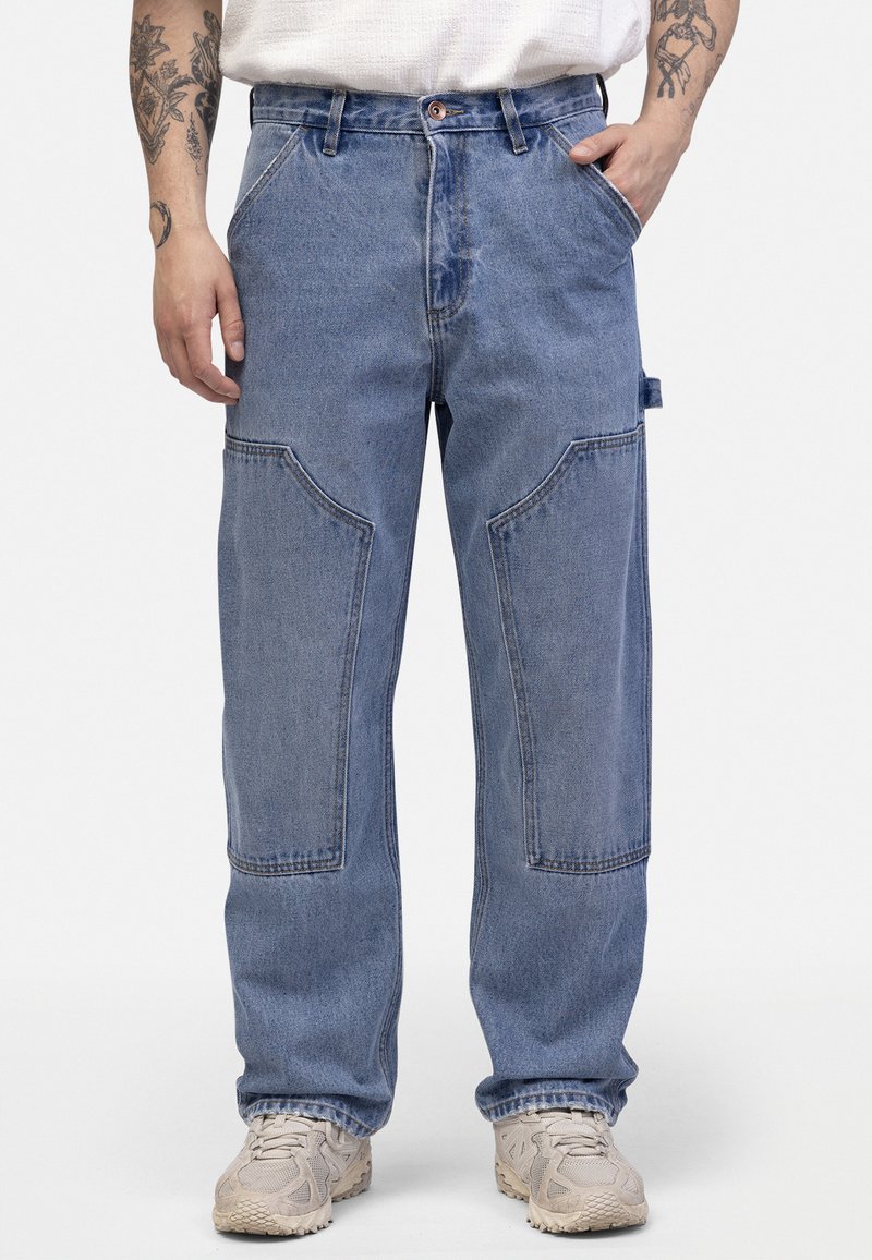 Pantalons en denim bleu clair à coupe décontractée, dotés de grandes poches avant arrondies et de coutures visibles. Portés avec des chaussures beiges.