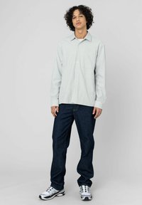 Chemise à boutons rayée en vert clair et blanc, associée à un jean en denim bleu foncé. Le modèle porte des chaussures de sport argentées avec des accents noirs.