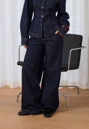 Personne portant une veste en jean foncée et un jean à jambes larges debout à côté d'une chaise de bureau noire sur un sol en bois.