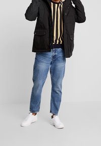 Chaqueta negra acolchada con bolsillos frontales sobre un suéter a rayas en azul marino y amarillo, combinada con jeans de mezclilla azul clara y zapatillas blancas.