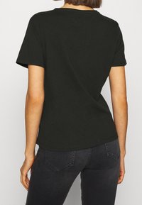 Woman wearing a plain black short-sleeve t-shirt and dark gray jeans, shown from the back against a neutral background.