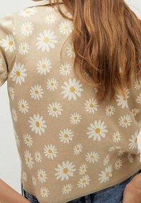 Beżowy sweter z dzianiny z wzorem stokrotek w białym i żółtym kolorze. Wzór jest reliefowy, a fason krótki z krótkimi rękawami.