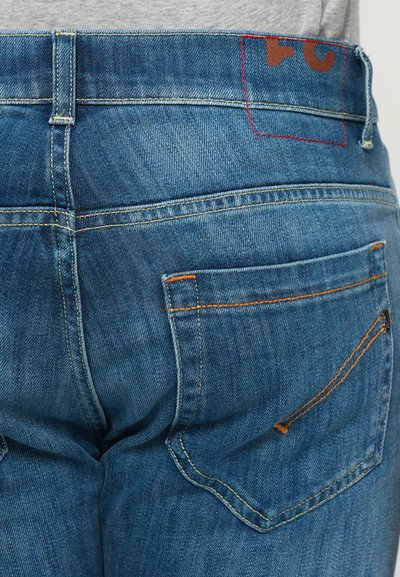 Gros plan de la poche arrière et de la taille d'un jean en denim bleu avec des coutures orange et un logo marron partiel sur la taille.