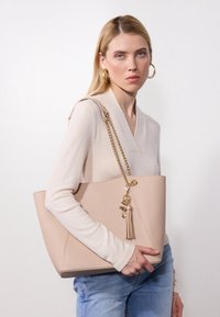 Beige leather tote bag with a textured finish, chain and faux-leather accents, featuring a tassel charm. Model holds bag against a light backdrop.