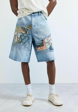 Muž nosiaci svetlomodré rifľové šortky do kolien s farebnými záplatami a švami, v kombinácii s bielymi ponožkami a krémovobielymi teniskami.