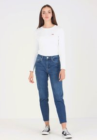 Weißes Langarmhemd mit kleinem rotem Logo, kombiniert mit blauen hochgeschnittenen Jeans und Schwarz-Weißen Sneakers, stehend vor einem schlichten Hintergrund.