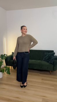 Μπεζ πλεκτό πουλόβερ με ριμπ ύφανση συνδυασμένο με σκούρο μπλε τζιν παντελόνι με φαρδύ πόδι, μαύρη τσάντα χειρός και γόβες με μυτερή μύτη, πάνω σε φόντο πράσινου καναπέ.