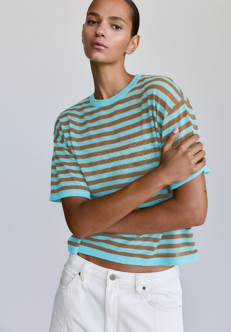 Jeune femme aux cheveux tirés en arrière, portant une chemise à manches courtes turquoise et marron rayée et un pantalon blanc, posant avec un bras croisé.