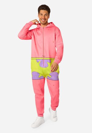 Homme souriant, portant un ensemble à capuche zippé rose et vert avec des taches violettes, debout devant un fond blanc.