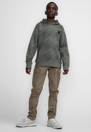 Felpa verde a motivi con un logo, abbinata a pantaloni cargo beige con molte tasche e sneakers di colore chiaro.