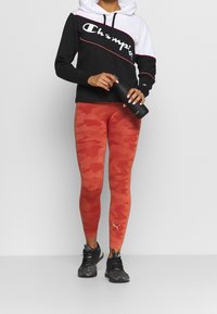 Svart och vit hoodie med logotyp, röda camo leggings, grå sneakers, håller en svart vattenflaska. Har randig design och elastiskt material.